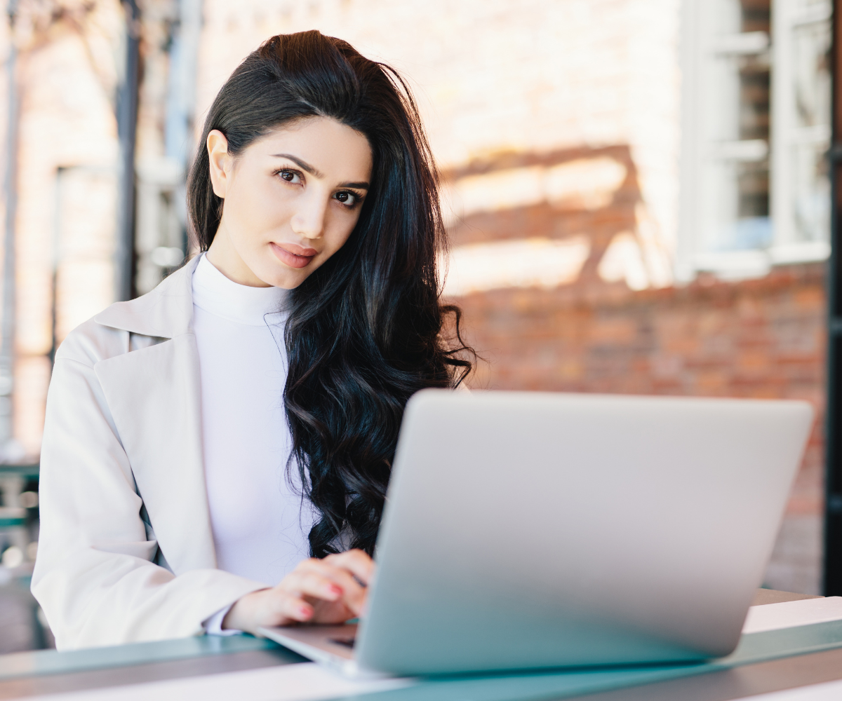 Mulher com cabelo longo e escuro, usando um blazer claro, trabalhando em um laptop em um ambiente moderno, representando serviços de consultoria e suporte em TI.
