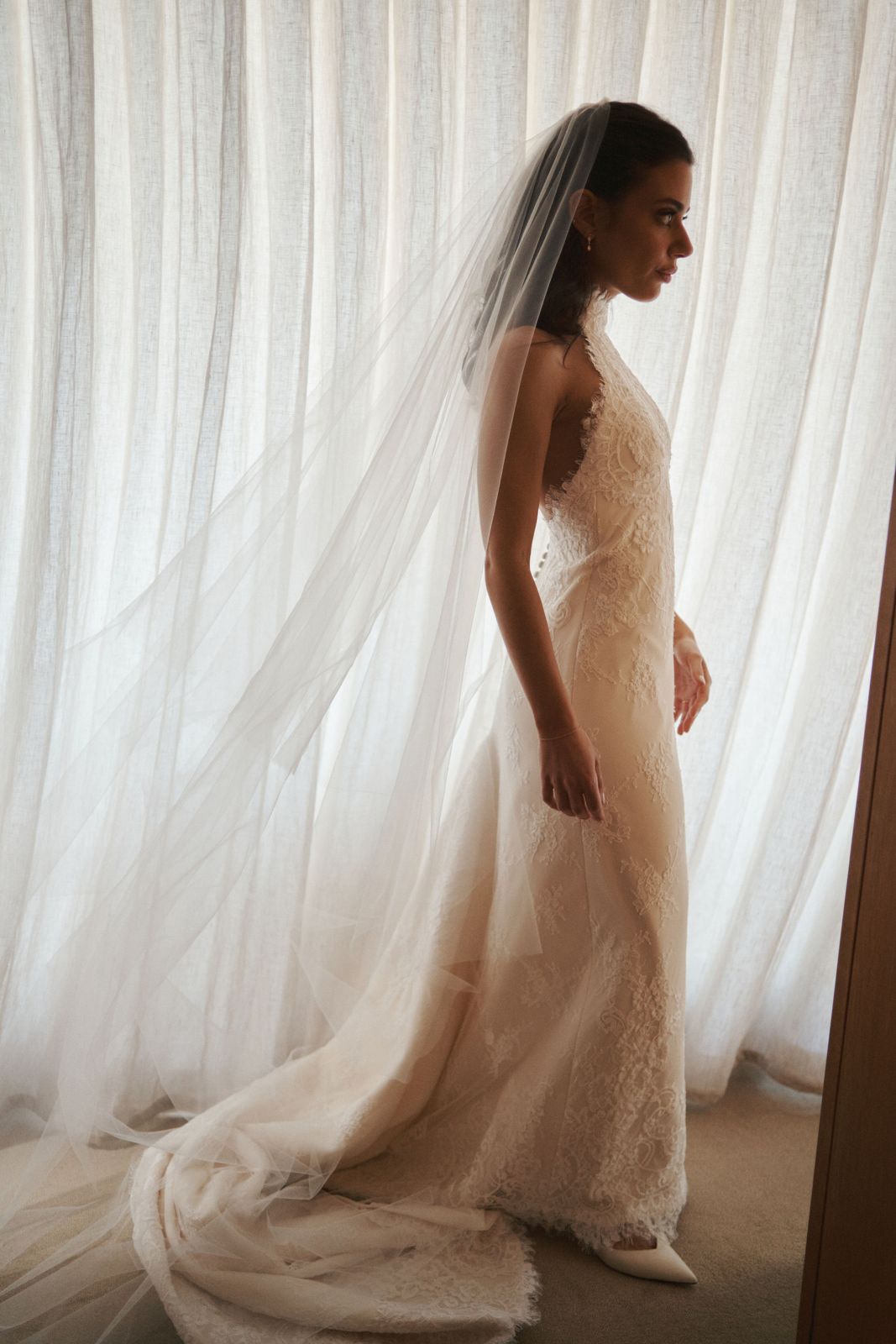 Real bride Jessie in a lace gown and cathedral veil by a sheer curtain