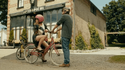 GIF: Alexis (Schitts Creek) is learning to ride a bike. Mutt is standing in front of the bike and holding the handlebars. The chain is off and Alexis is just peddling