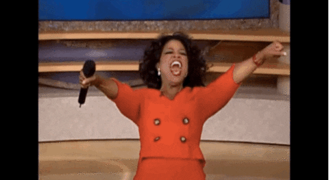 GIF: Oprah is joyfully yelling and pointing at the audience with both hands after giving everyone a car. 
