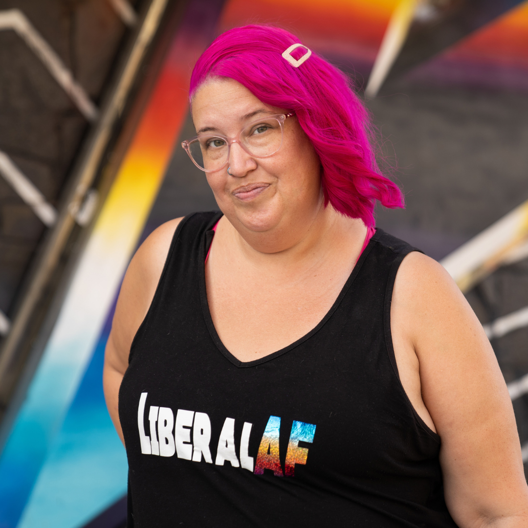 Meg is standing and smiling at the camera. She wears a "LiberalAF" tank top and has hot pink hair.