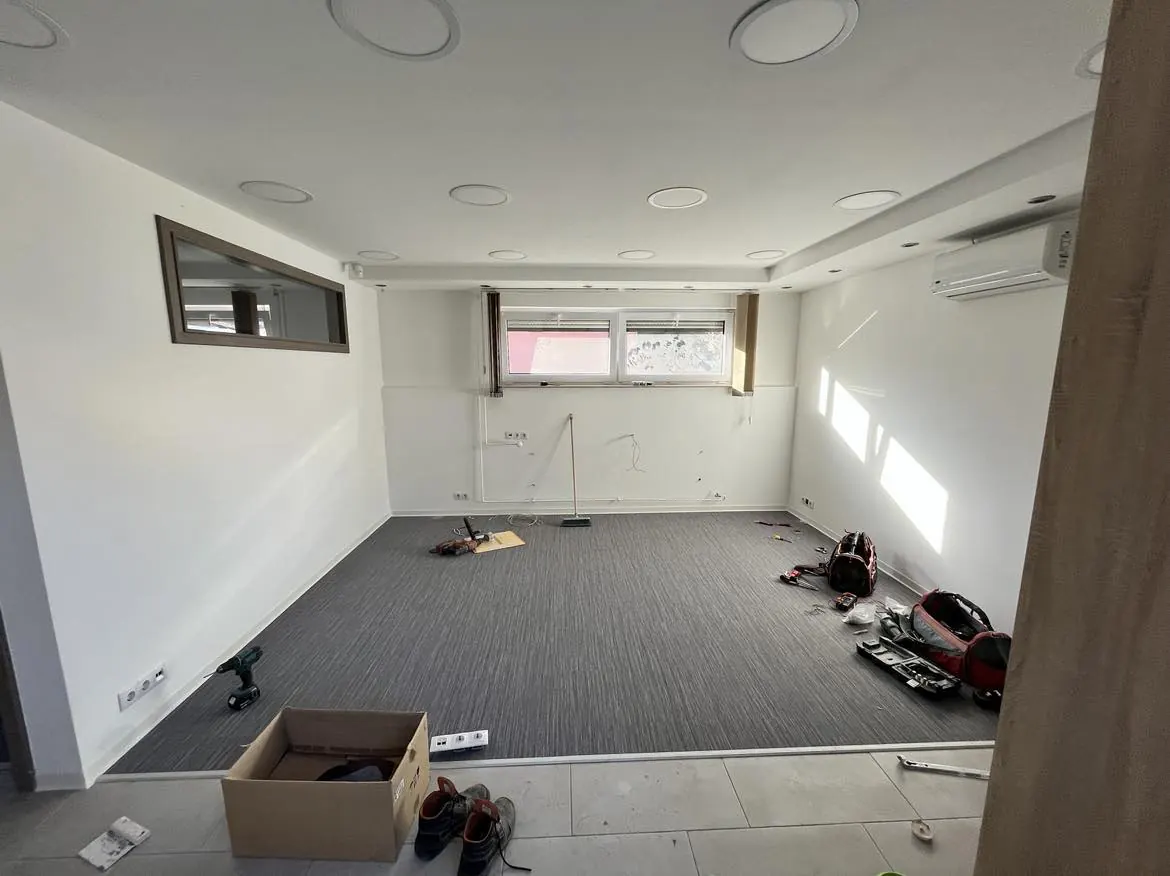 A képen egy frissen felújított, világos irodai tér látható, amely Győr városában található. A falak fehérre festettek, a padlón szürke szőnyeg található. Az ablakok mellett természetes fény áramlik be, míg a mennyezeten modern, kör alakú világítótestek helyezkednek el. Az irodában néhány szerszám és munkaeszköz látható, ami a Farkas-Konstrukt Kft. Generálkivitelezés tevékenységére utal.