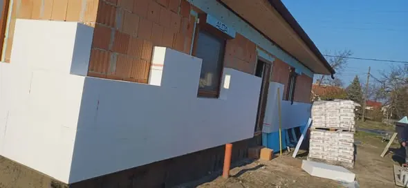 A képen a Farkas-Konstrukt Kft. hőszigetelési munkálatai láthatók Győr egyik épületén. Az épület falán fehér hőszigetelő panelek kerülnek elhelyezésre, míg a háttérben látható a részleges téglafal. Az építkezési területen anyagok, köztük tégla és egyéb építőanyagok találhatók, amelyek a generálkivitelezés folyamatát segítik elő. Az időjárás napos, tiszta égbolttal.