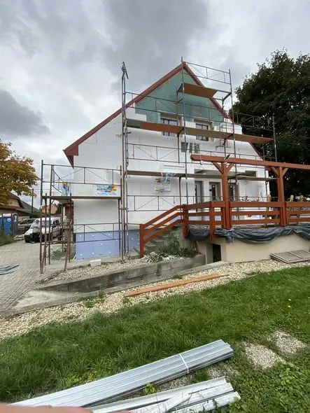 A képen egy építkezés látható Győrben, ahol a Farkas-Konstrukt Kft. generálkivitelezés szolgáltatását végzik. Az épület homlokzata részben felújítás alatt áll, állványzattal körbevéve, míg a kertben építőipari anyagok, például acéllemez látható. A ház kulcsrakész kivitelezésének előkészületei zajlanak, ami a modern építkezési trendeknek megfelelően történik.