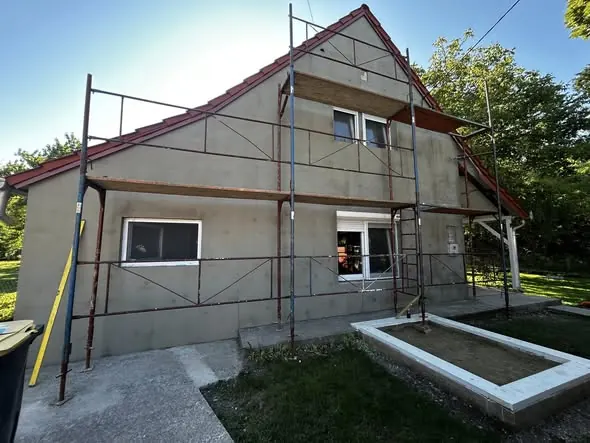 A képen egy folyamatban lévő építkezés látható, ahol a Farkas-Konstrukt Kft. generálkivitelezés szolgáltatásait végzik. Az épület külső falai látszanak, amelyek már vakolt állapotban vannak, és az épület előtt állványzat helyezkedik el. Az építkezés Győr városában zajlik, és a kulcsrakész kivitelezés során az épület felújítása folyamatban van. A zöld terület és a napfényes időjárás kellemes hátteret ad az építkezéshez.