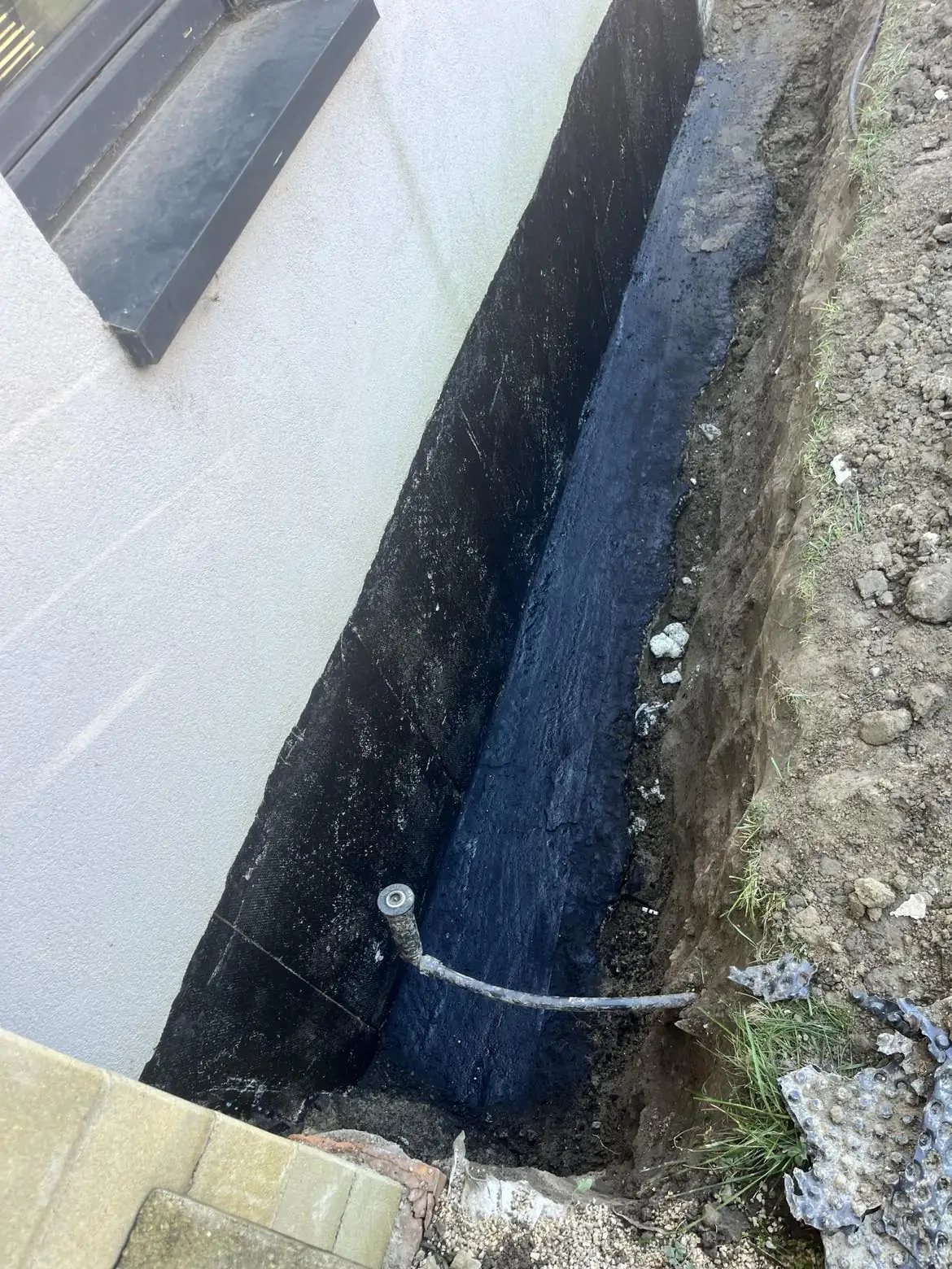 A képen egy építkezés látható, ahol egy ház alapjának vízszigetelését végzik. A földbe ásott területen fekete vízszigetelő anyag látható, amely megakadályozza a víz bejutását. A háttérben a ház falának részletei és egy vízelvezető cső is megfigyelhető. A Farkas-Konstrukt Kft. Generálkivitelezés szolgáltatásaival Győr városában biztosítja az építkezések szakszerű kivitelezését.