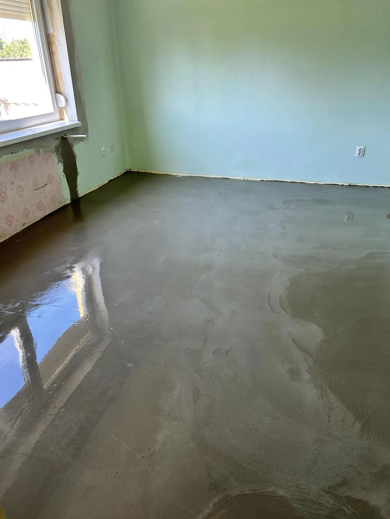 A Kőszegen található Farkas-Konstrukt Kft. generálkivitelezési munkájaként bemutatott szoba belső terében frissen elkészült, sima betonpadló látható. A falak világos zöld színűek, és az ablakon keresztül természetes fény áramlik be. A padló felülete csillogó, egyenletes, ami a szakmai kivitelezés magas színvonalát tükrözi.