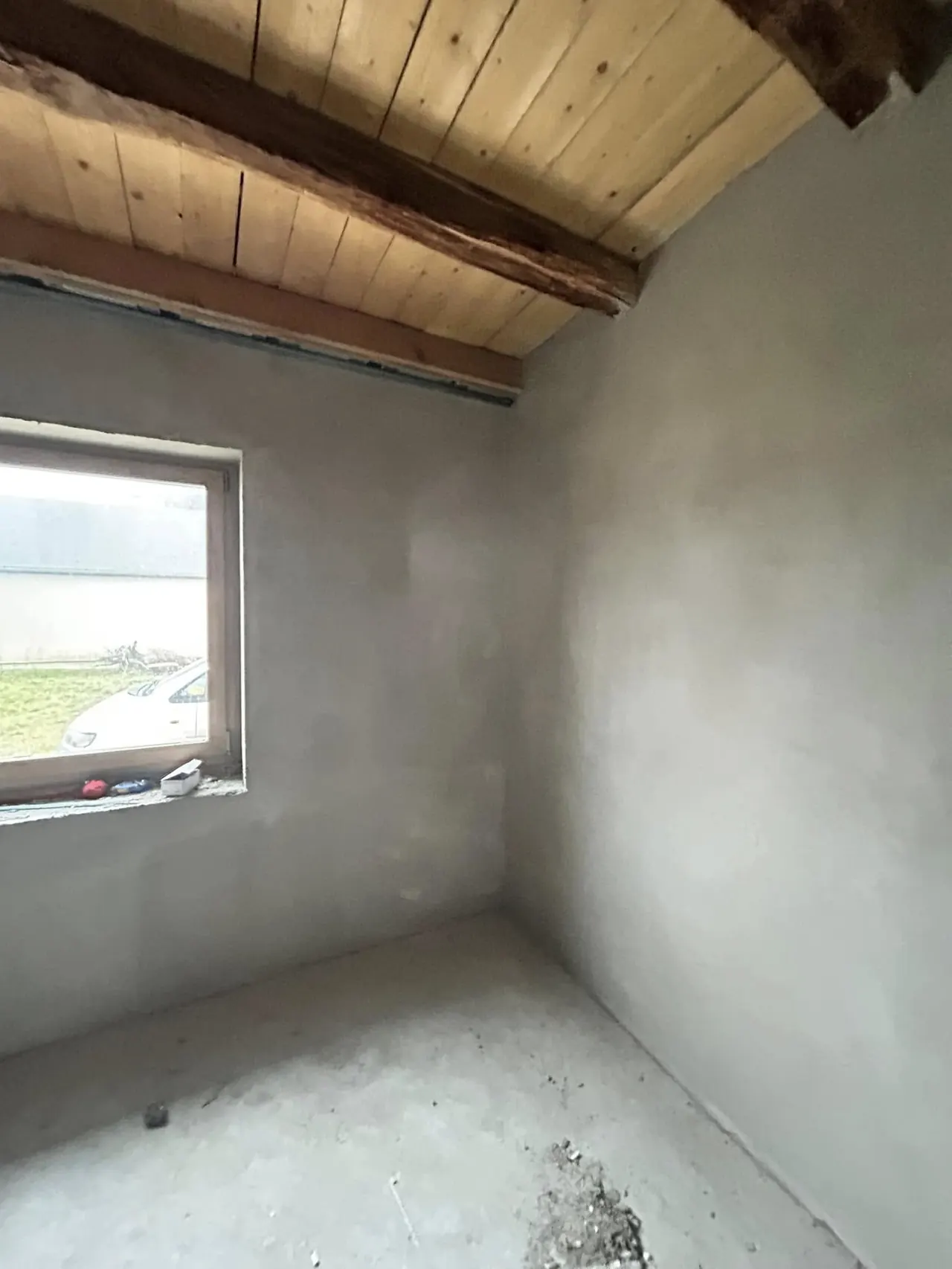 A képen egy frissen kivitelezett belső tér látható, ahol a falakat simára vakolták, és a mennyezet fából készült gerendákkal díszített. Az ablakon keresztül természetes fény árad be, amely megvilágítja a még befejezés előtt álló helyiséget. A Farkas-Konstrukt Kft. által végzett generálkivitelezés Győr városában zajlik, amely a modern építkezési megoldásokat tükrözi.