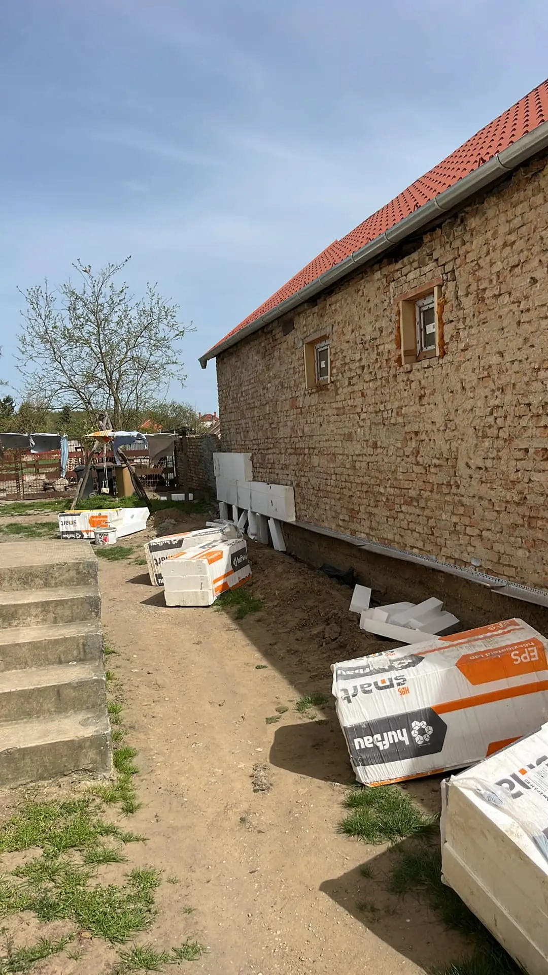 A képen a Farkas-Konstrukt Kft. generálkivitelezéshez szükséges építőanyagok láthatók, amelyek éppen a győri építkezés területén találhatók. A fal mellett fehér szigetelőanyagok vannak elhelyezve, míg a háttérben egy nem befejezett épület és zöld növényzet látható. A terület rendezett, és a lépcsők a ház bejáratához vezetnek.