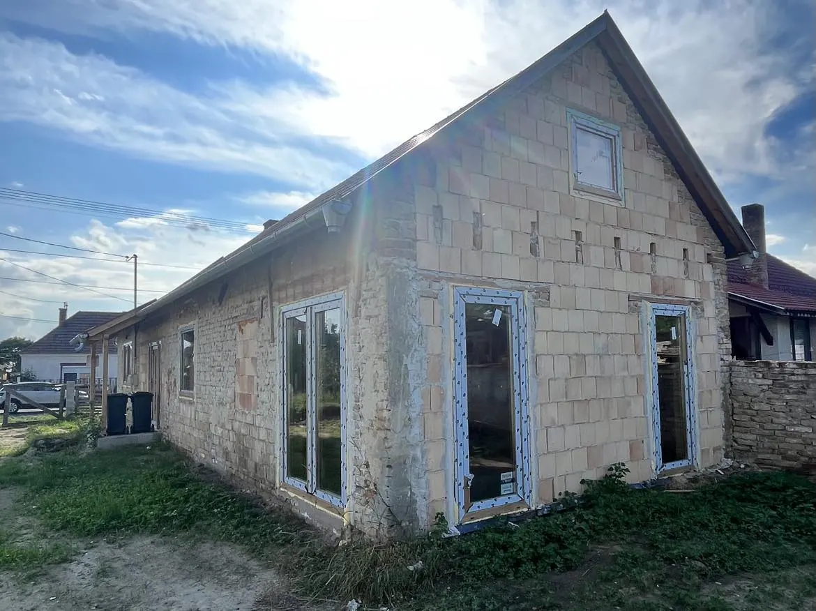 A képen egy Sárváron található építkezés látható, ahol a Farkas-Konstrukt Kft. generálkivitelezési munkálatai zajlanak. A ház homlokzata részben kész, láthatók az ablakkeretek, valamint a falon a szigetelés előkészítése folyamatban van. A napfény szépen megvilágítja az épületet, ami a jövőbeli otthonok megvalósítását jelzi. Az építkezés környezete rendezett és jól karbantartott.