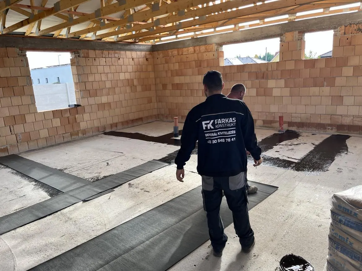 A képen a Farkas-Konstrukt Kft. munkatársai láthatók, amint a győri építkezésen a padló szigetelését készítik elő. Az épület belső terében a falak már készen állnak, és a munkafolyamat során a munkások precízen helyezik el a szigetelő anyagokat. A Generálkivitelezés területén szerzett tapasztalatukkal biztosítják a kiváló minőséget.