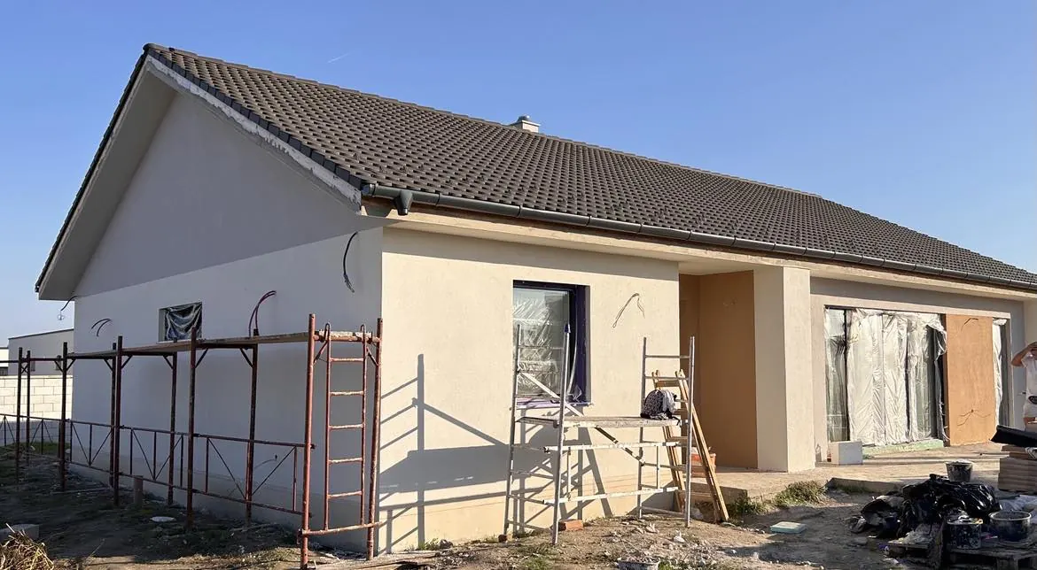 Újonnan épült ház látható Győrben, ahol a Farkas-Konstrukt Kft. generálkivitelezési szolgáltatásait végzik. Az épület falai simára vannak vakolva, és az ablakok körüli munkálatok zajlanak. Az építkezés körül állványzat található, amelyen munkások dolgoznak, miközben a terület rendezése alatt áll. A képen a szép idő és a világos égbolt is látható.