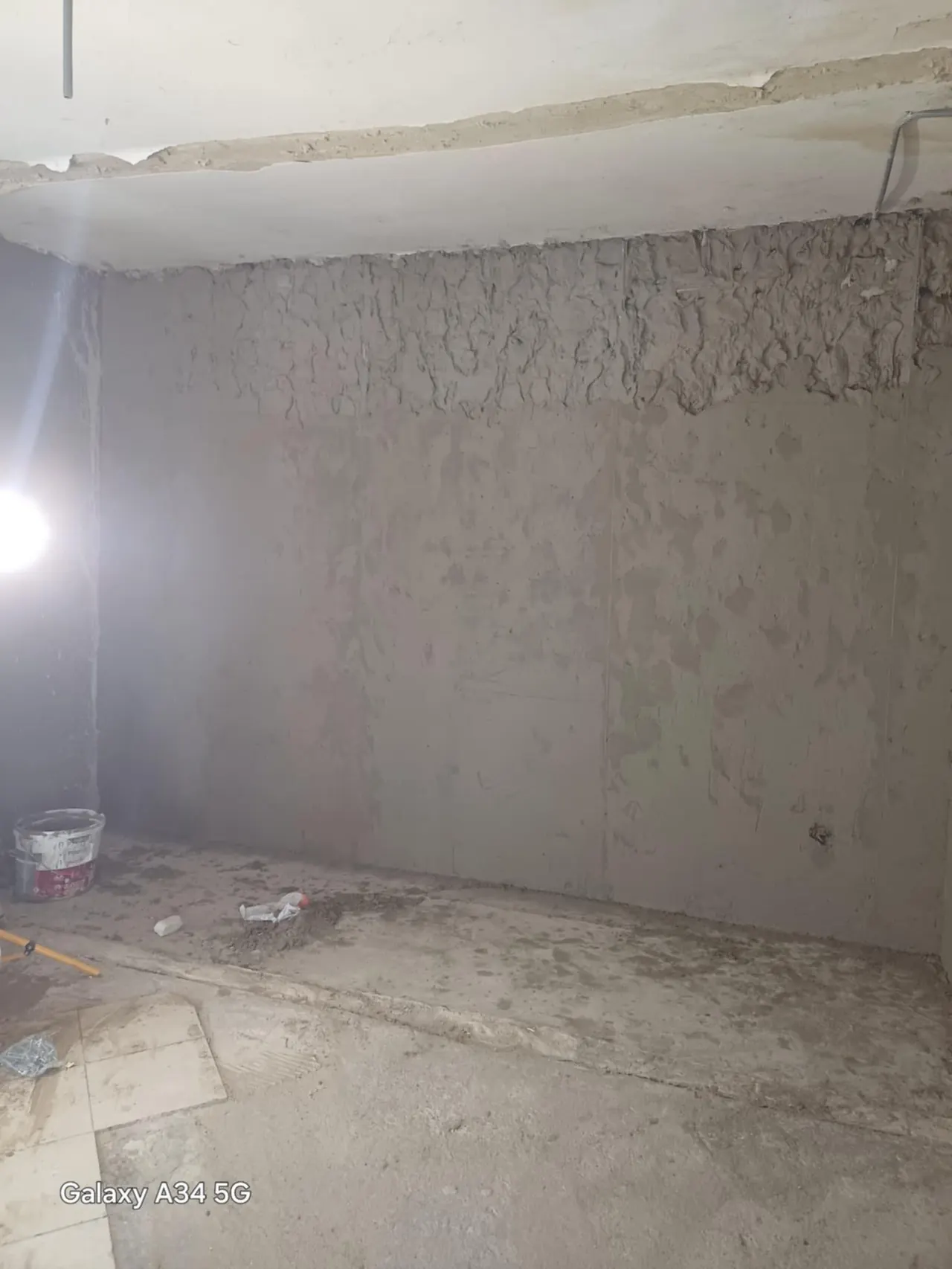 A képen egy lakásfelújítási folyamat látható, ahol a falak előkészítése zajlik. A szoba még nem befejezett állapotban van, a falak durva, simítatlan felülete látható, és a padlón különböző építőanyagok találhatóak. A háttérben egy világítótest segítségével világítanak, ami fokozza a munkaterület fényviszonyait. A Farkas-Konstrukt Kft. Generálkivitelezés szolgáltatása Győr városában zajlik.