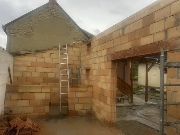 Fertőd városában a Farkas-Konstrukt Kft. Generálkivitelezés munkálatai láthatók, ahol egy épület falának építése zajlik. A képen a téglafal, létra, és a felújítás alatt álló ház részletei láthatók. Az építkezés környezete rendezett, különböző építőanyagokkal, mint például tégla és vakolat, a háttérben pedig a ház régi falának maradványai is megfigyelhetők.