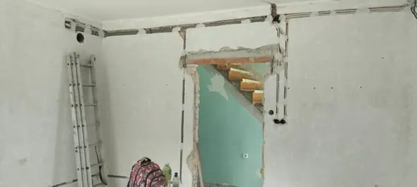 A képen egy lakásfelújítás folyamatban lévő munkaterülete látható Győr városában. A falon látható repedések és a nyitott ajtókeret a generálkivitelezés során végzett bontási munkákat jelzi. A háttérben lévő létra és a táska a munkavégzéshez szükséges eszközöket tükrözi. A Farkas-Konstrukt Kft. szakértelme egyértelműen megjelenik a felújítás előkészítésében.