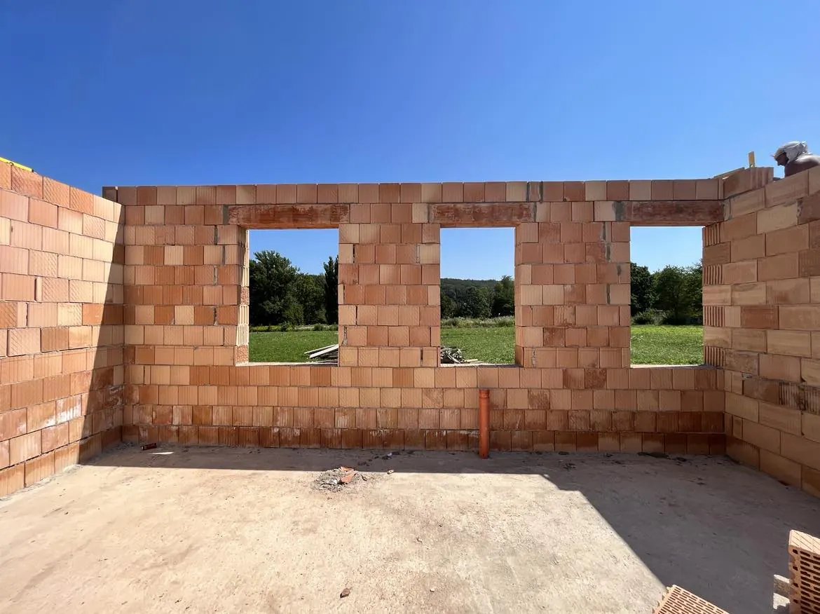 A képen látható egy építkezési terület Győr városában, ahol a Farkas-Konstrukt Kft. generálkivitelezési munkálatai zajlanak. A falon három ablaknyílás látható, amelyeket téglaépítkezés jellemez. A háttérben zöld fák és növényzet látható, a tiszta kék ég alatt. Az építkezés előrehaladott állapotban van, jól tükrözve a vállalat szakmai színvonalát.