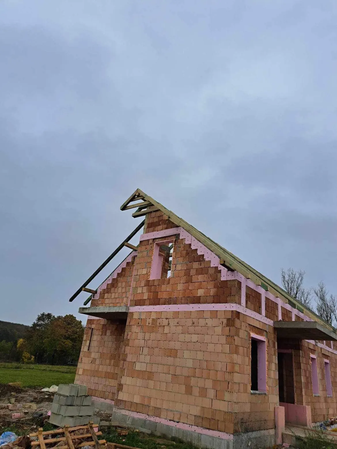 Győr városában a Farkas-Konstrukt Kft. Generálkivitelezés szolgáltatásának keretében egy épülő ház látható. Az épület falai téglából készülnek, és a tetőszerkezet részben már kialakításra került. A képen a ház formája és a nyílások jól kivehetőek, a háttérben pedig fák és szürke égbolt látható.