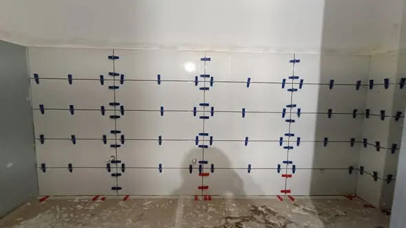 A képen látható egy frissen burkolt fal, amelyen fehér csempe található, kék és piros csempefülek segítségével rögzítve. A burkolás precíz, egyenletes elrendezés figyelhető meg, amely a Farkas-Konstrukt Kft. generálkivitelezési munkáját tükrözi. A helyszín Várpalota, ahol a csempe munkálatok kiváló minőségben készültek.