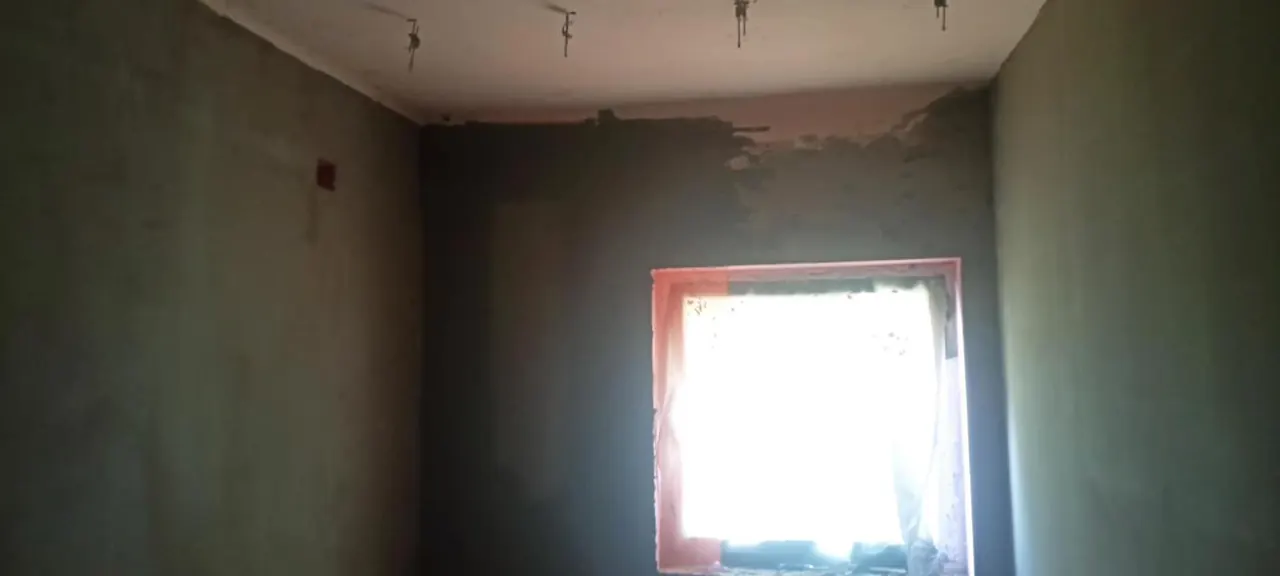 A képen egy nemrégiben felújított szoba látható, amelynek falai frissen glettelt, szürke színű bevonattal vannak ellátva. A szoba egyik oldalán egy nagy ablak található, amely mögött természetes fény árad be, világosabbá téve a teret. Az ablakkeret vörös színű, ami kontrasztot ad a falak színével. Az Esztergomban működő Farkas-Konstrukt Kft. generálkivitelezési tevékenységének részeként a szoba felújítása folyamatban van.