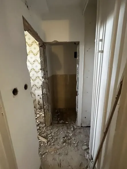 A képen egy felújítás alatt álló belső tér látható, ahol a falak mellett törmelék és por található. A folyosón egy nyílás látható, amely egy másik helyiségbe vezet, a falak részben le vannak bontva. A háttérben csempe látszik, ami a fürdőszoba vagy konyha területére utal. Ez a munka a Farkas-Konstrukt Kft. generálkivitelezési projektje Tatabányán zajlik.