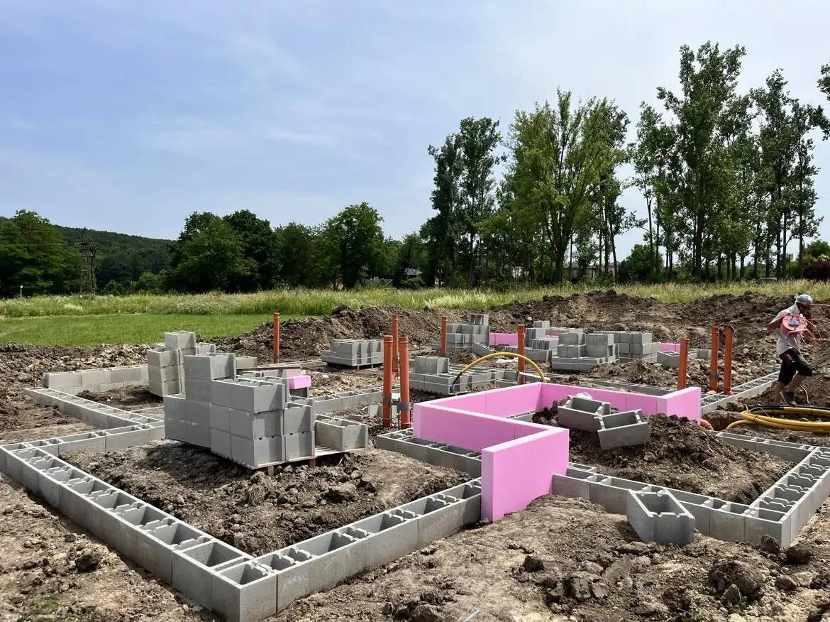 A Farkas-Konstrukt Kft. generálkivitelezési projektje látható Győr közelében, ahol éppen az alapozás folyik. A képen szürke betonblokkokból készült alapkeret és élénk rózsaszín elemek láthatók, amelyek a jövőbeli épület határait jelölik. A munkálatok közepette a zöld fák és a tiszta égbolt is megfigyelhető, ami kellemes környezetet teremt az építkezés számára.
