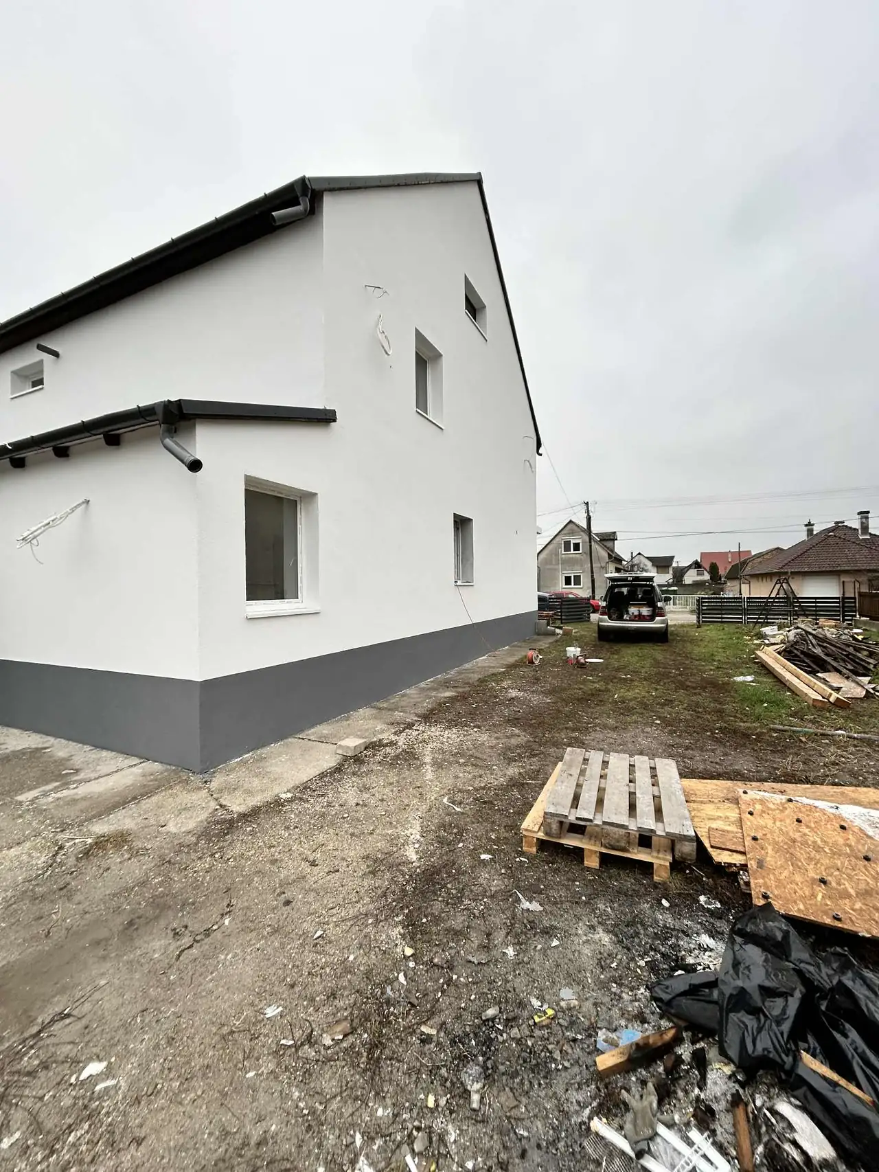 A képen egy frissen festett, fehér színű ház látható, amelynek oldala és tetője jól láthatóan új állapotú. A ház mellett építési törmelék és fából készült raklapok találhatók. A háttérben más épületek is látszanak, ami a győri környezetet tükrözi. A Farkas-Konstrukt Kft. generálkivitelezési munkálatai során készült el ez a projekt, amely Győr városában található.