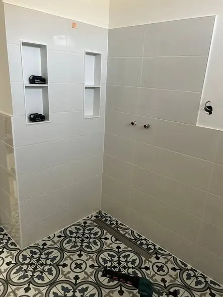 A képen egy frissen felújított fürdőszoba látható, amely Győr városában készült a Farkas-Konstrukt Kft. generálkivitelezésének keretében. Az új, világos csempék modern megjelenést kölcsönöznek a helyiségnek, míg a mintás padlóburkolat egyedi karaktert ad. A falba épített polcok és a vízcsapok már a végső simításokhoz készülnek, ami a szakértelmet és a precizitást tükrözi.