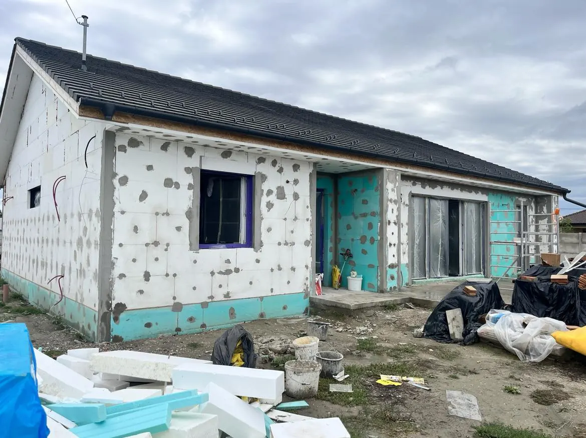 Győr városában, a Farkas-Konstrukt Kft. generálkivitelezési projektjén készült fotón egy épülő ház látható, amelynek külseje részben kész, de még befejezésre vár. Az épület falai fehér szigetelőanyaggal vannak borítva, míg az ablakok keretei kék színűek. A területen építőanyagok és szerszámok találhatók, ami az aktív munkálatokat tükrözi.