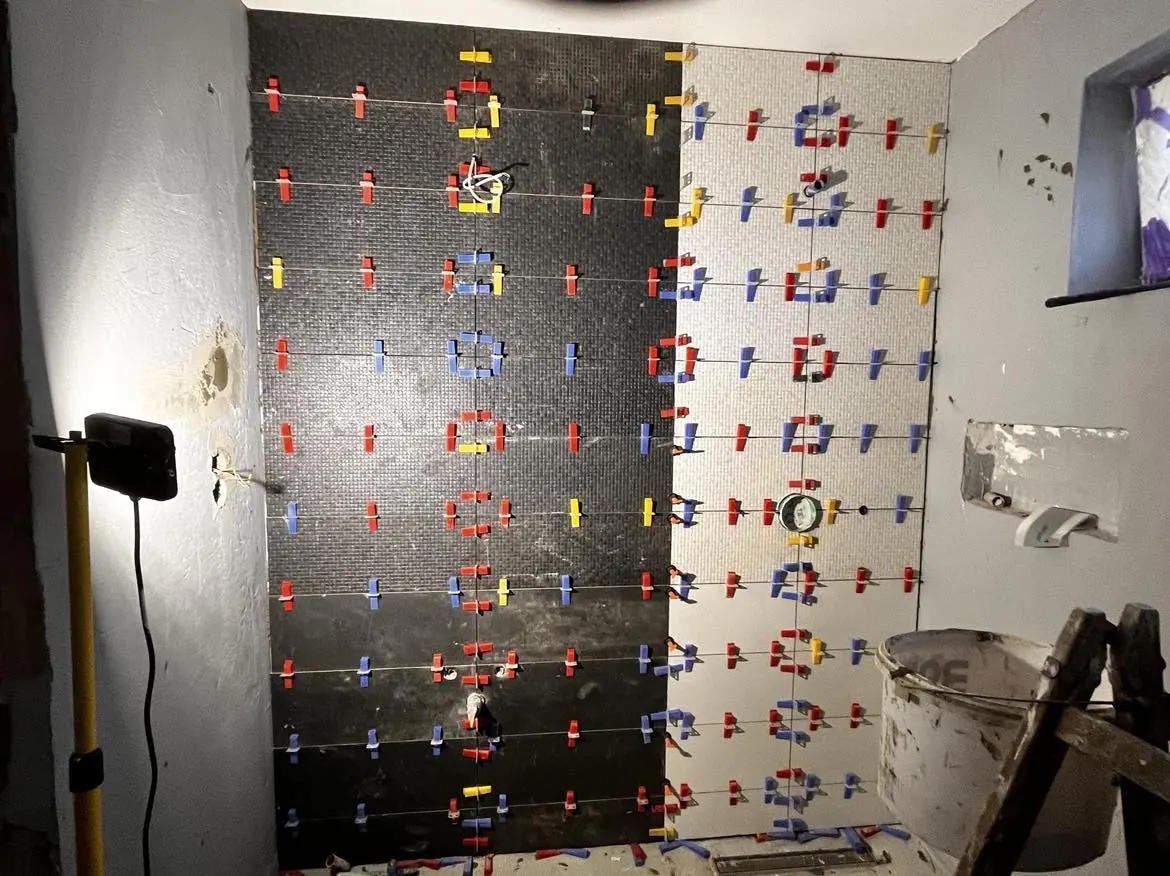 A képen egy félkész csempeburkolat látható, amely fekete és szürke csempékből áll, sárga és piros csempefugázó klipszekkel rögzítve. A háttérben egy lámpa világít, és a fal mellett egy festékes vödör található. A helyszín Kapuvár, ahol a Farkas-Konstrukt Kft. generálkivitelezési munkálatait végzi.