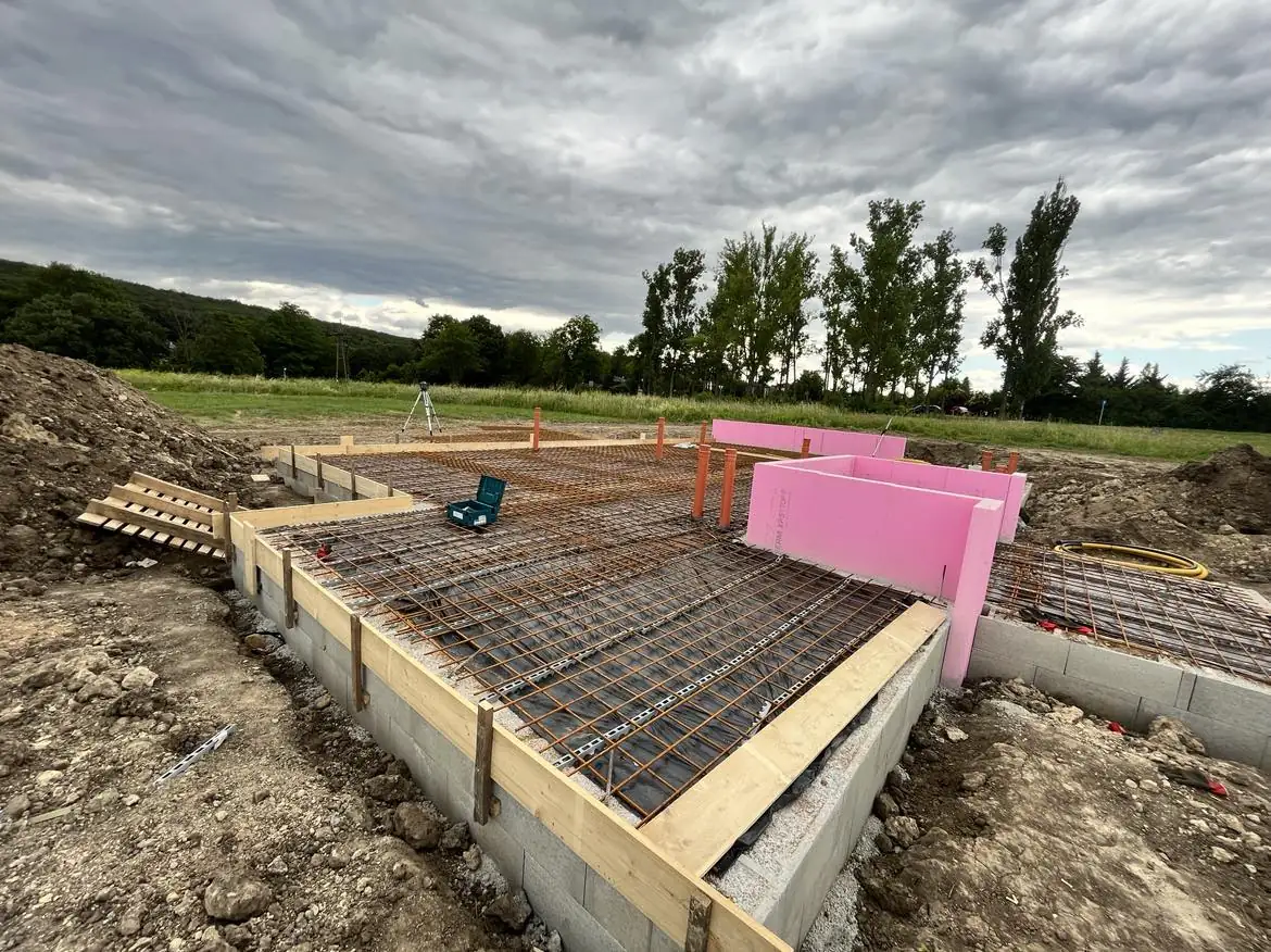 Győr városában, a Farkas-Konstrukt Kft. generálkivitelezési projektjén készülő alapozás látható. Az építkezés során jól látható a beton alap, amelyen acél szerkezet és hőszigetelő elemek találhatók. A háttérben zöld fák és egy szürke égbolt díszíti a tájat, ami a munkaterület nyugodt környezetét hangsúlyozza.