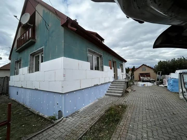 A képen egy Tata városában található épület látható, amelynek generálkivitelezése a Farkas-Konstrukt Kft. által folyik. Az épület oldalfala szigetelve van, fehér polisztirol lapokkal, és a ház előtt lépcső vezet a bejárathoz. A háttérben más épületek is láthatók, míg az építkezési területen különböző anyagok találhatóak, amelyek a kivitelezéshez szükségesek.