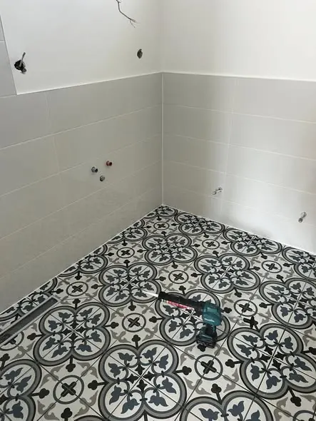 A képen egy frissen felújított fürdőszoba látható, ahol a falakat világos csempék borítják, míg a padlón mintás, fekete-fehér járólap található. A falakon vízcsapok előkészítése látható, ami a Generálkivitelezés folyamatát tükrözi. A kép Győr városában készült, ahol a Farkas-Konstrukt Kft. szakértelme érvényesül a modern fürdőszobák kialakításában.