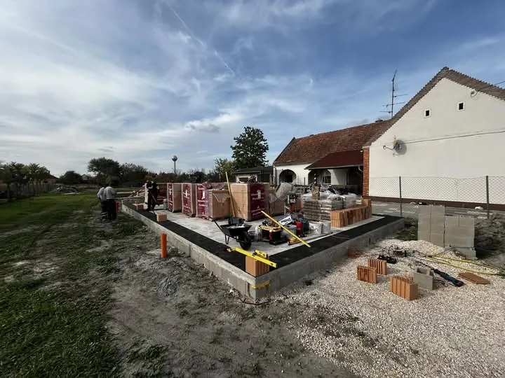 A képen egy építkezés látható Bük városában, ahol a Farkas-Konstrukt Kft. generálkivitelezési munkálatait végzik. Az építkezés területén különböző építőanyagok, mint téglák és betonblokkok találhatók, valamint munkások, akik az alapozást készítik elő. A háttérben egy ház és fák láthatók, a felhők pedig szép időt jeleznek.