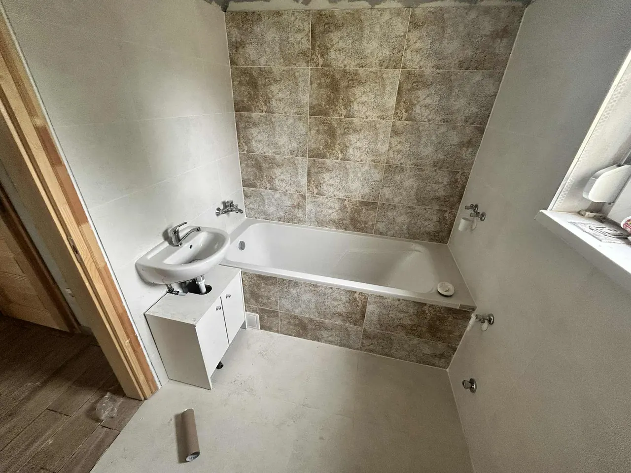 A képen egy fürdőszoba felújítása látható, ahol a falakat modern, barna és bézs árnyalatú csempe borítja. Az új kád és a mosdó helye már ki van alakítva, a padló pedig sima, világos színű burkolattal van ellátva. A Farkas-Konstrukt Kft. szakemberei Győrben végzik a generálkivitelezést, hogy a fürdőszoba modern és praktikus legyen, a munka még folyamatban van.