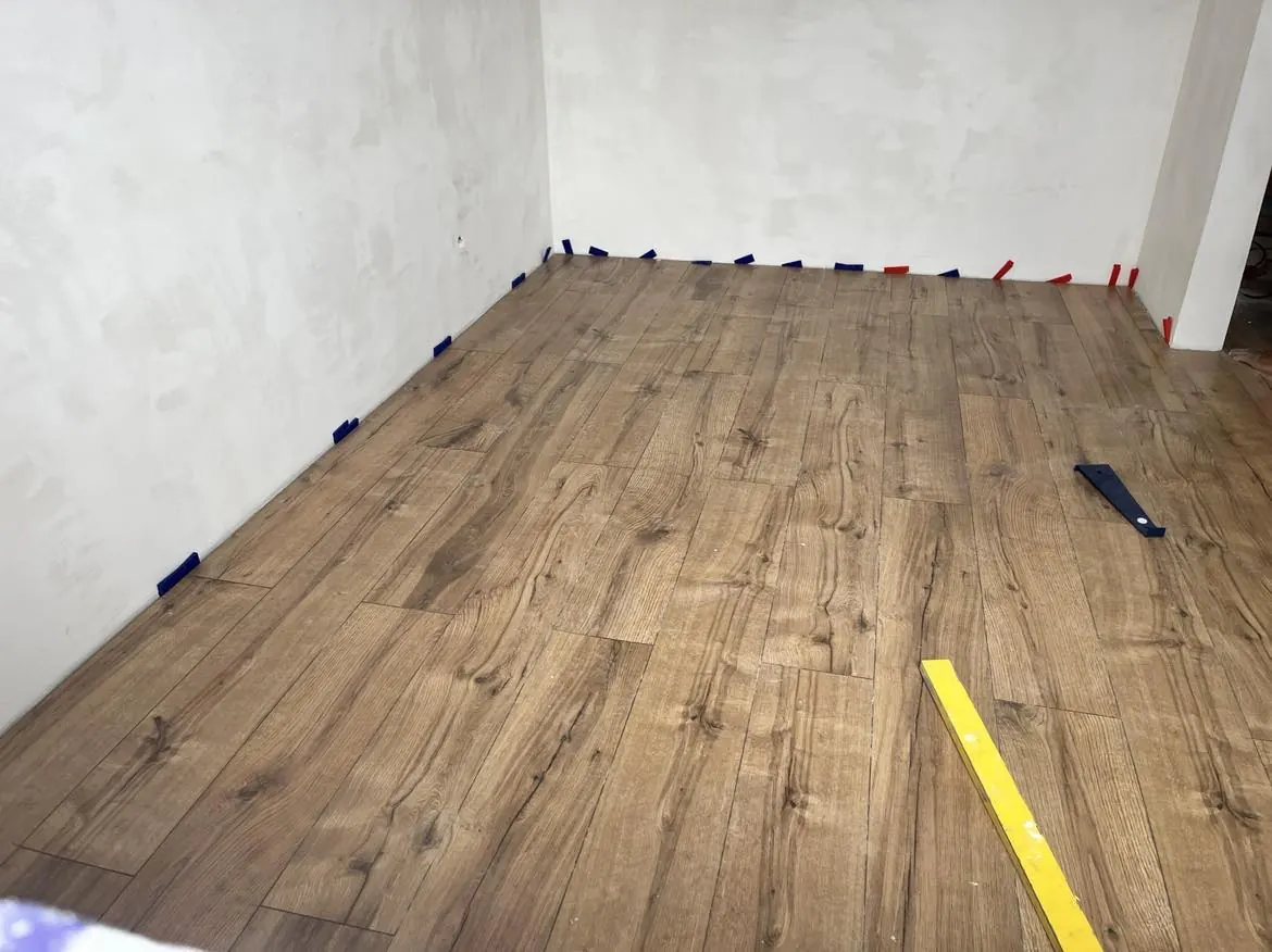 Az image egy frissen burkolt helyiséget ábrázol, ahol világosbarna laminált padló található. A padló szélén különböző színű jelölőszalagok láthatók, amelyek segítik a burkolási munkálatok pontos végrehajtását. A falak simára vannak glettelve, és a háttérben a munka kezdeti fázisa látható. A Farkas-Konstrukt Kft. Generálkivitelezés szolgáltatásait Győr városában nyújtja.