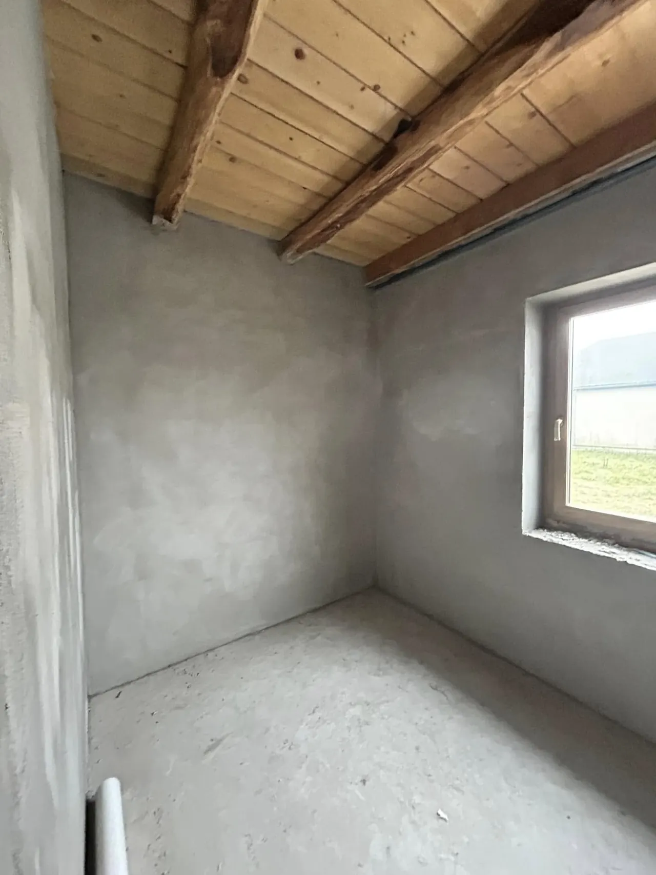 A képen egy frissen épített belső tér látható, amelynek falai simára vakoltak, és a mennyezet fából készült gerendákkal van díszítve. Az ablakon keresztül természetes fény áramlik be, amely világosabbá teszi a helyiséget. A tér Győr városában található, és a Farkas-Konstrukt Kft. generálkivitelezési munkálatainak része.