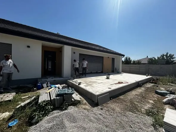 Új építésű ház területe látható Győr városában, ahol a Farkas-Konstrukt Kft. Generálkivitelezés szolgáltatásait végzi. A képen munkások dolgoznak a teraszon, amely már le van burkolva, és a háttérben egy modern ház áll. A területen építőanyagok és szerszámok találhatóak, a napfény világítja meg a munkaterületet.