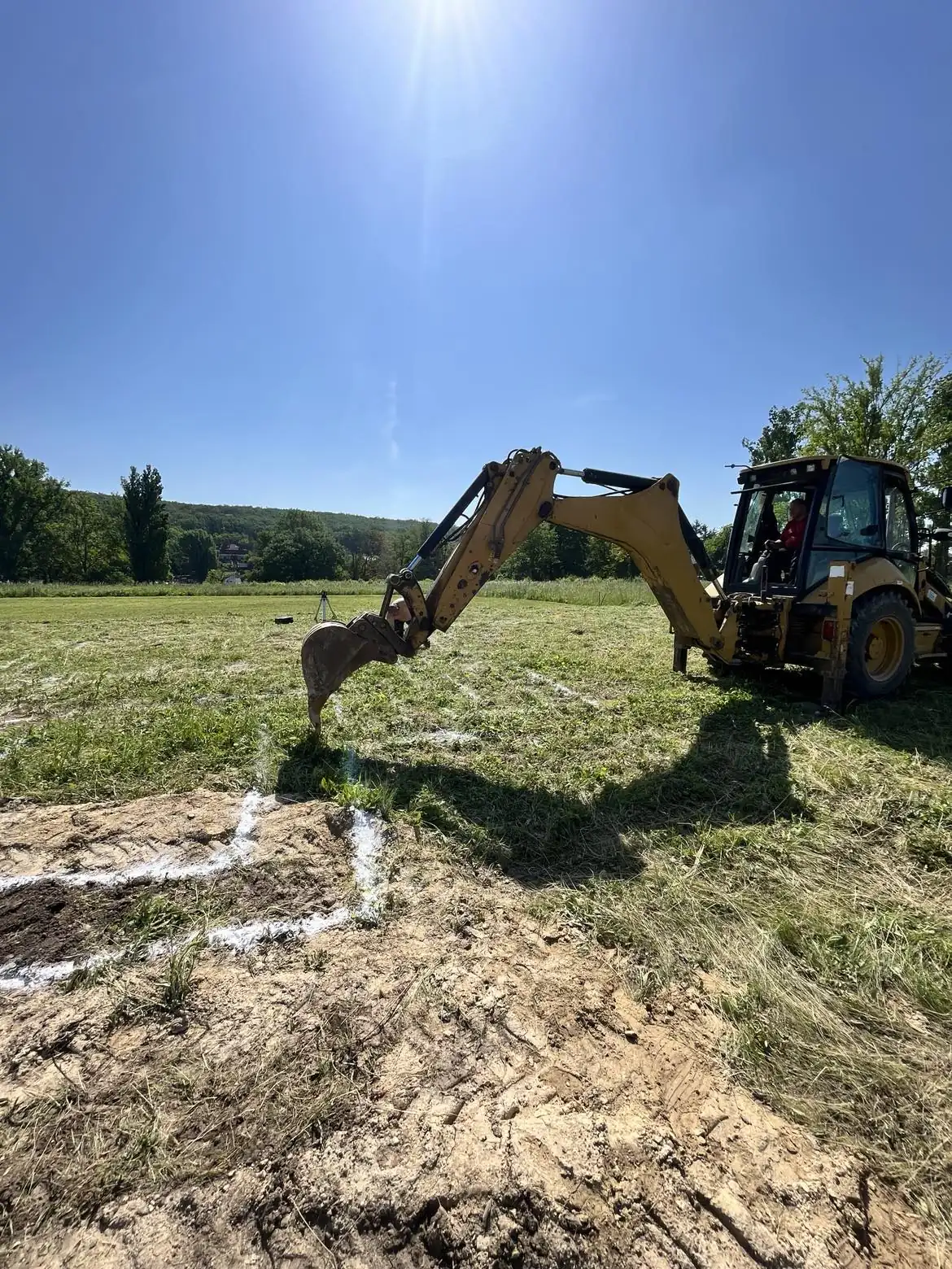 Egy napfényes napon, Kapuvár területén a Farkas-Konstrukt Kft. Generálkivitelezés csapata munkába állt. A képen egy sárga kotrógép látható, amely éppen egy építkezéshez szükséges földmunkát végez. A háttérben zöld fák és dombok láthatók, amelyek szép tájat alkotnak, míg a munkagép előtt frissen megmunkált terület található.