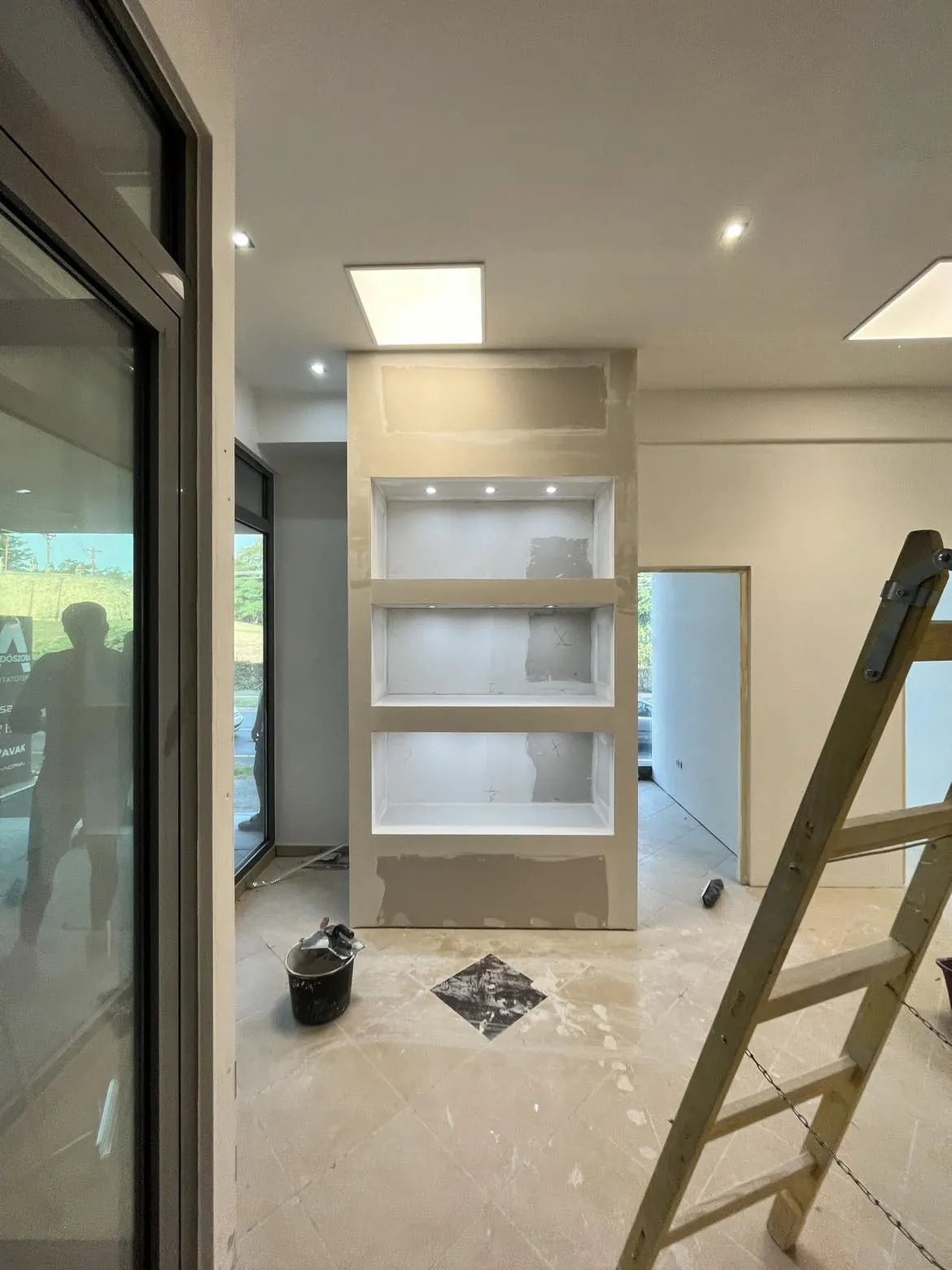 A képen egy frissen felújított belső tér látható, amelyben a falon világító polcok kerültek kialakításra. A szoba padlója burkolt, és a falak simára vannak vakolva. A háttérben egy létra áll, amely a Generálkivitelezés folyamatát jelzi. A képet Sopronban készítették, ahol a Farkas-Konstrukt Kft. szakemberei dolgoznak a projekt kivitelezésén.