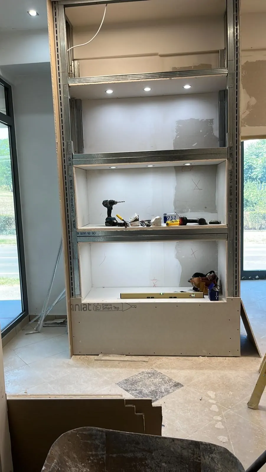 A képen egy folyamatban lévő lakásfelújítás látható, ahol a Farkas-Konstrukt Kft. generálkivitelezés szolgáltatását végzik Győrben. Az építkezés során egy falra szerelt polcrendszert alakítanak ki, amely világítással van ellátva. Az asztalnál különböző szerszámok, például fúró és építőanyagok találhatók, amelyek a felújításhoz szükségesek.