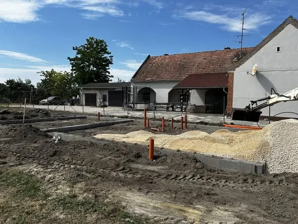 A képen a Farkas-Konstrukt Kft. generálkivitelezési munkálatai láthatók Győrben. Az építkezés területén homok és kavics halmok találhatóak, valamint már megkezdődött az alapozás, ahol narancssárga PVC csövek állnak ki a földből. A háttérben egy régi ház látható, amelynek felújítása folyamatban van. Az időjárás napos, kék égbolttal.