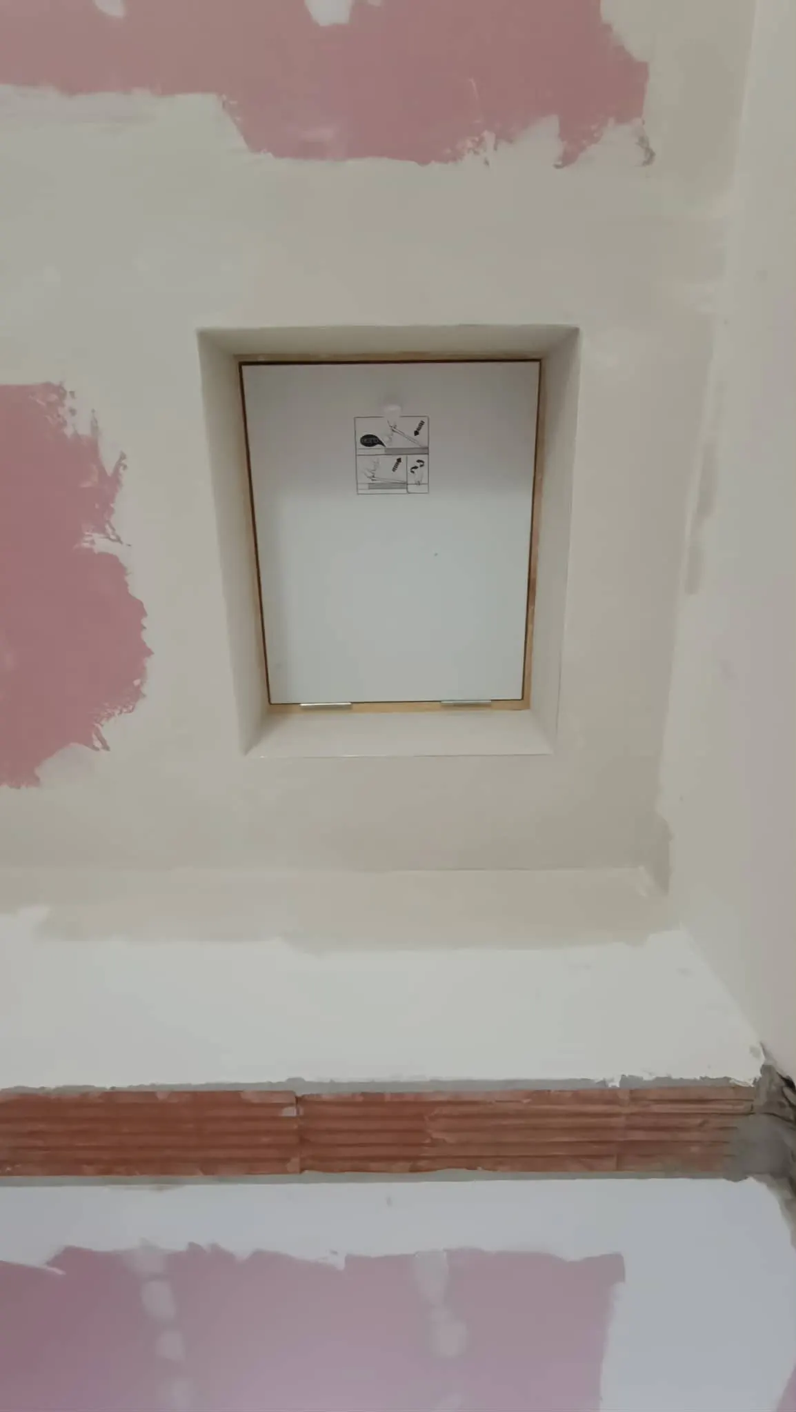 A képen egy burkolási folyamat látható, ahol frissen glettelt fal található. A fal közepén egy fehér kerettel ellátott vaknyílás látható, amely mellett rózsaszín festékfoltok vannak. A háttérben a fal alsó részén cserép burkolat helyezkedik el. Az ilyen típusú generálkivitelezési munkák a Farkas-Konstrukt Kft. szakértelmét tükrözik Győr városában.