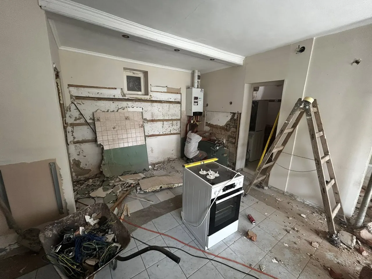 A képen egy felújítás alatt álló lakás látható, ahol a falak részben lebontva, a padló pedig építőanyagokkal és törmelékkel van tele. Az egyik falon látható egy konyhai sütő, míg a háttérben egy munkás dolgozik a vízvezeték szerelésén. Az egész terület rendetlen, ami a Generálkivitelezés folyamatát tükrözi. A város neve, Győr, jól illeszkedik a Farkas-Konstrukt Kft. tevékenységéhez.