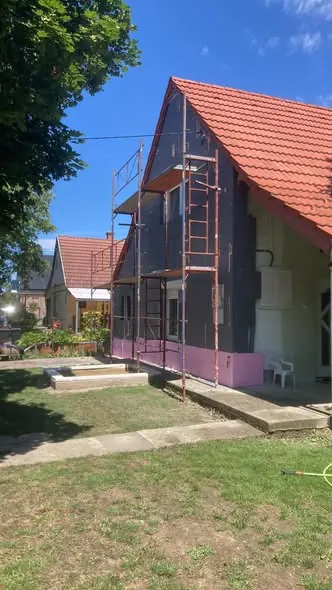 A képen egy építkezés látható, ahol a Farkas-Konstrukt Kft. szakemberei dolgoznak egy ház kulcsrakész kivitelezésén Győr városában. Az épület homlokzata éppen felújítás alatt áll, állványzat került felállításra, és a környező terület rendezett. A háttérben lakóházak is láthatók, amelyek a generálkivitelezés folyamatát illusztrálják.