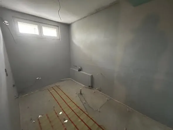 A képen egy frissen felújított szoba látható, amelynek falai világosszürkére vannak festve. A padlón látható a fűtési rendszer előkészítése, ahogy a csövek elhelyezésére szolgáló szigetelőanyag is. Az ablakok fényesek, és sok természetes fényt engednek be. A Farkas-Konstrukt Kft. által végzett generálkivitelezés nyomai jól láthatók Győr városában.