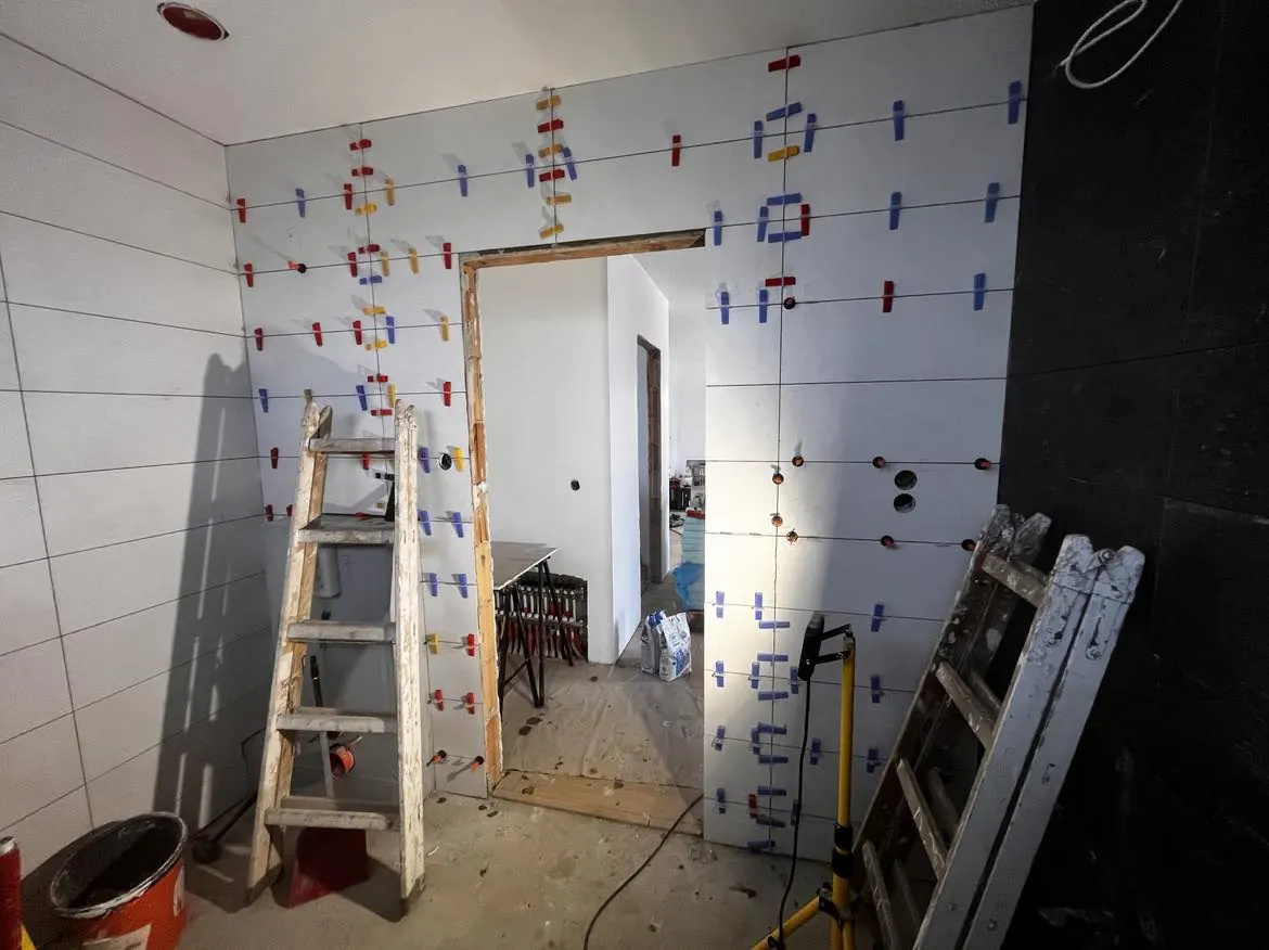 Győr városában, a Farkas-Konstrukt Kft. generálkivitelezési projektje keretében készült felvétel. A képen egy frissen burkolt fal látható, amelyen csempefektető klipszek vannak elhelyezve. A háttérben egy nyitott ajtókeret található, míg a földön csempeburkolási munkákhoz szükséges eszközök, például létra és fúró láthatók. A tér rendezett, de még folyamatban lévő kivitelezési munkálatok nyomait mutatja.