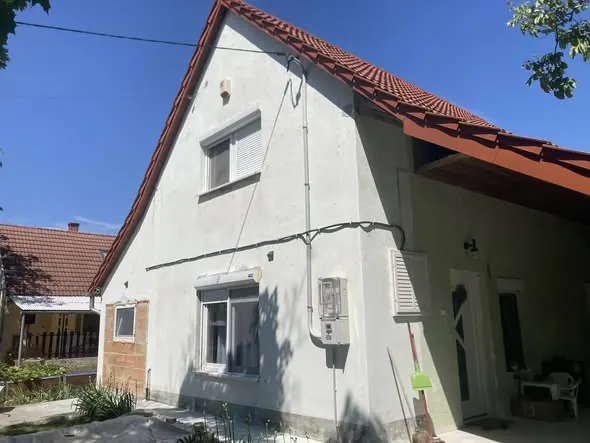 A képen egy frissen felújított ház látható Komárom városában, ahol a Farkas-Konstrukt Kft. Generálkivitelezés szolgáltatásait használták. A ház homlokzata világos színű, az ablakok előtt redőnyök találhatók. A tető cseréppel fedett, a kertben pedig zöld növények láthatók, ami barátságos atmoszférát kölcsönöz az ingatlan környezetének.