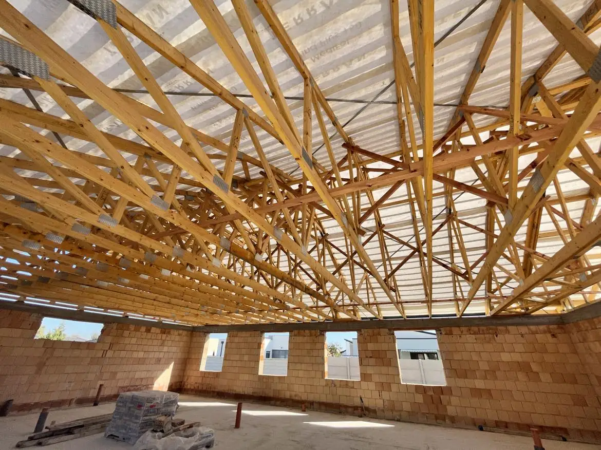 A képen a Farkas-Konstrukt Kft. generálkivitelezési projektje látható Győr városában. Az épület belső terében a fa tetőszerkezet jól megfigyelhető, amely szimmetrikusan elhelyezkedő gerendákból áll. A falak még nem készültek el, a padlón téglák és építőanyagok találhatók. A természetes fény átszűrődik a tetőn, világos légkört teremtve az építkezés során.