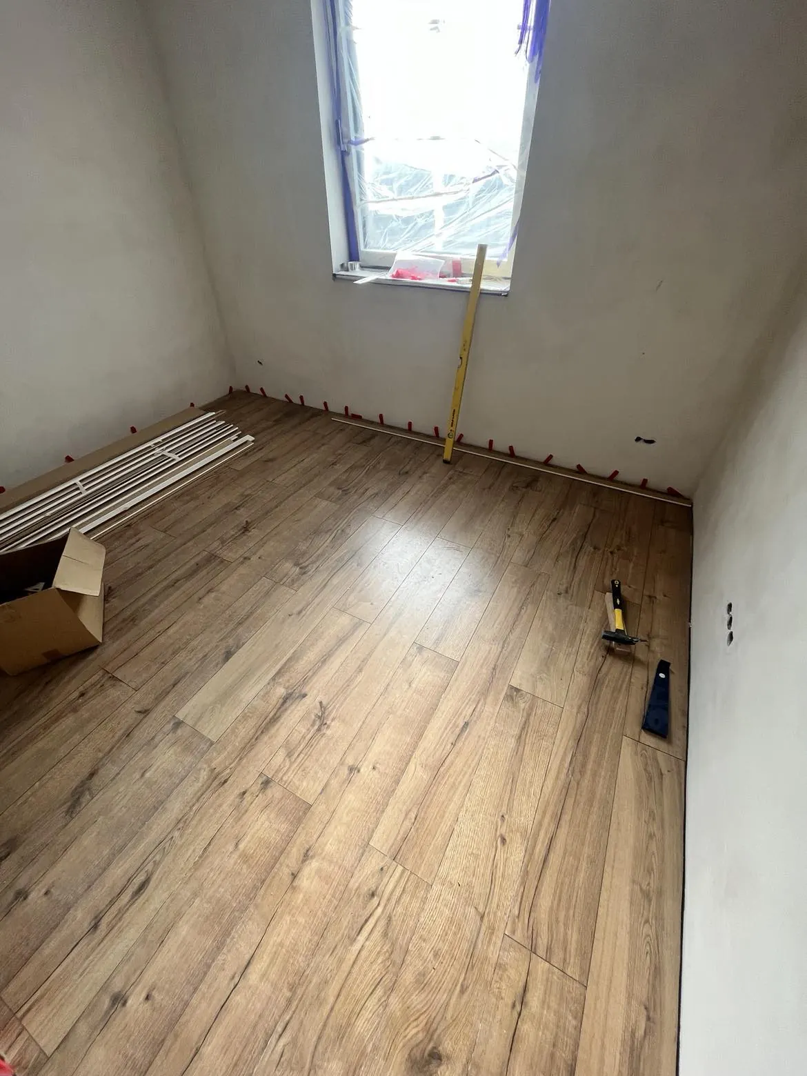 A képen egy frissen felújított szoba látható Kőszegen, amely a Farkas-Konstrukt Kft. Generálkivitelezés szolgáltatásának eredménye. A falak világos színűek, a padlózaton fa hatású laminált burkolat található. A szobában lévő eszközök, mint például egy létra és egy tűzőgép, a befejezés előtti munkálatokat mutatják. Az ablak előtt védőfólia található, ami a további munkákat segíti.