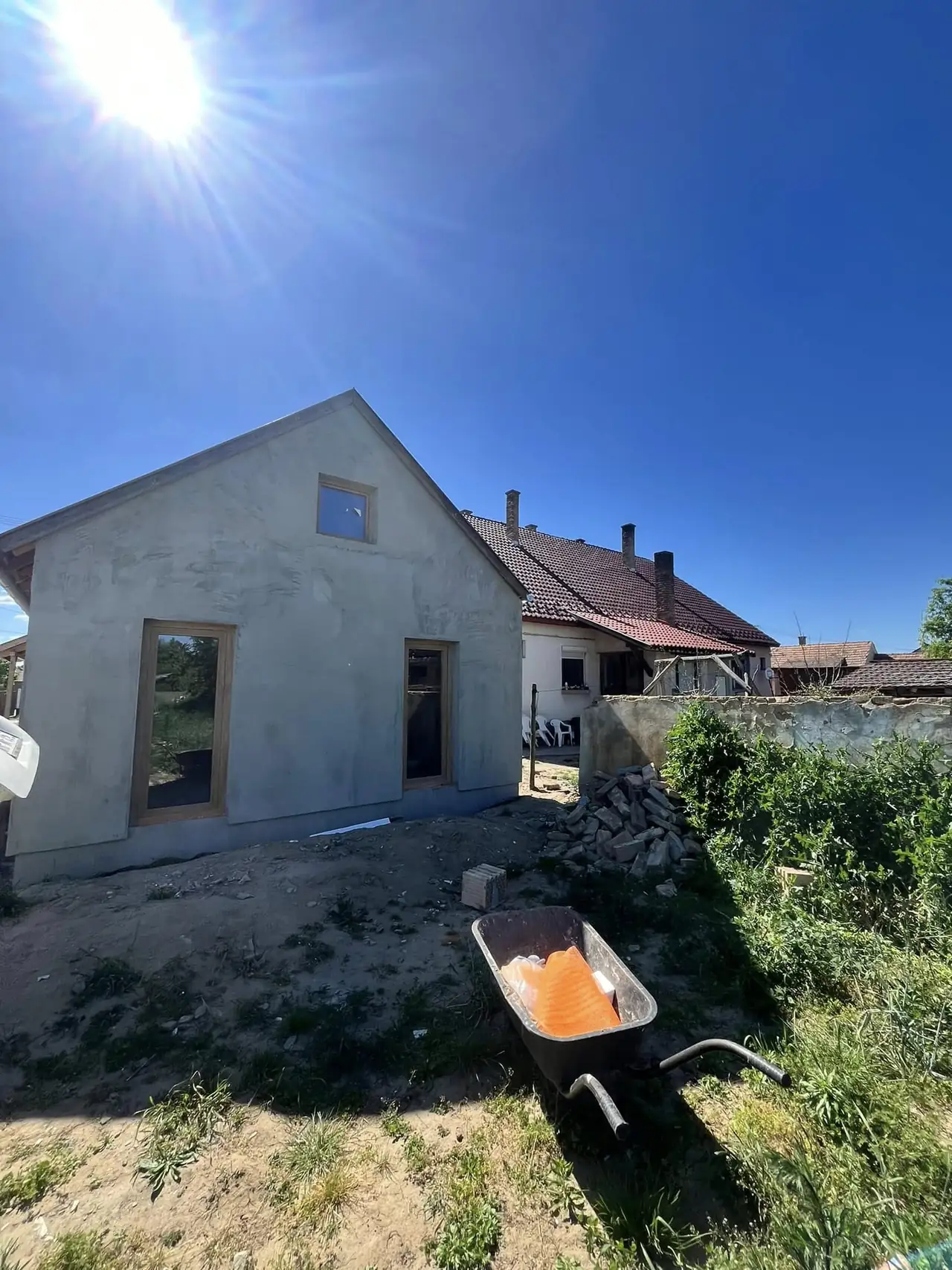 A képen egy építkezés látható Győr városában, ahol a Farkas-Konstrukt Kft. Generálkivitelezés tevékenysége zajlik. A frissen vakolt ház látható, amelynek ablakai már be vannak helyezve. A háttérben egy másik épület és egy kerékpáros talicska áll, tele homokkal, amely a munkálatokhoz szükséges. Az idő napos, tiszta égbolt alatt zajlik az építkezés.